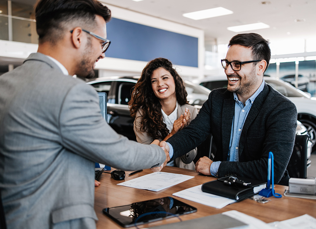 Insurance by Industry - View of a Smiling Business Man Shaking Hands with a Young Couple Buying a Car at a Car Dealership
