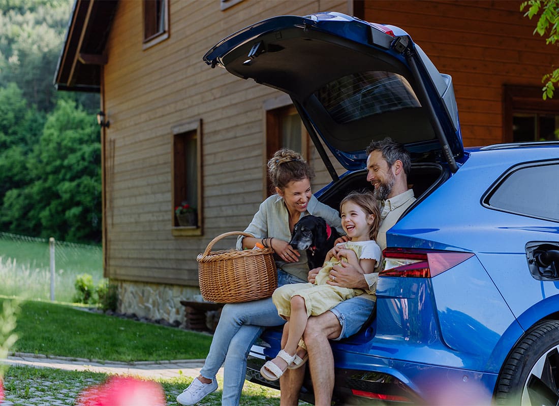 Personal Insurance - Portrait of a Cheerful Family with a Young Daughter Sitting in the Trunk of an SUV Next to Their House While its Charging